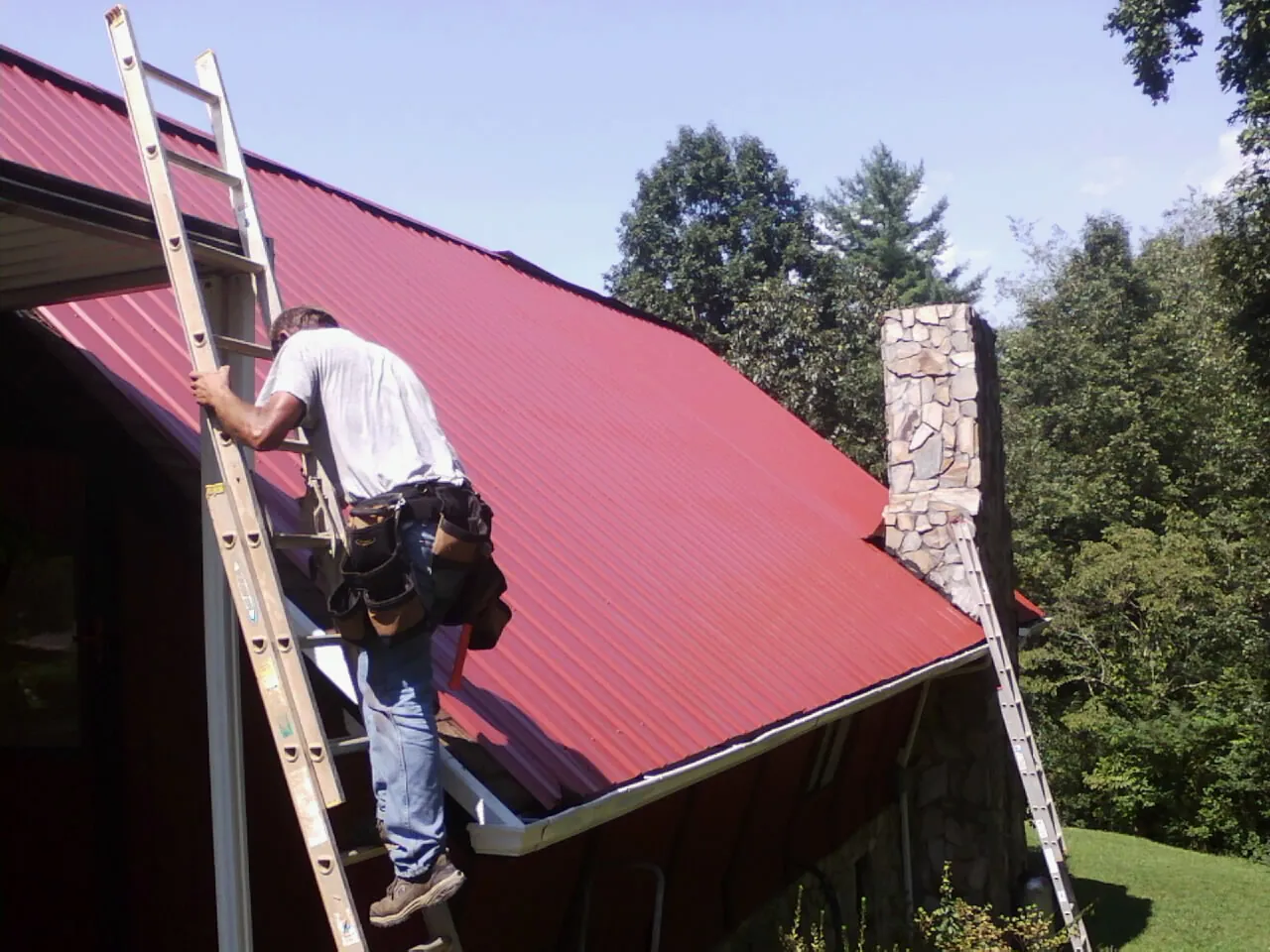Roofer on a ladder beside a red metal roof and stone chimney — Soffit & Fascia Repair by Roofing Brothers in Munday